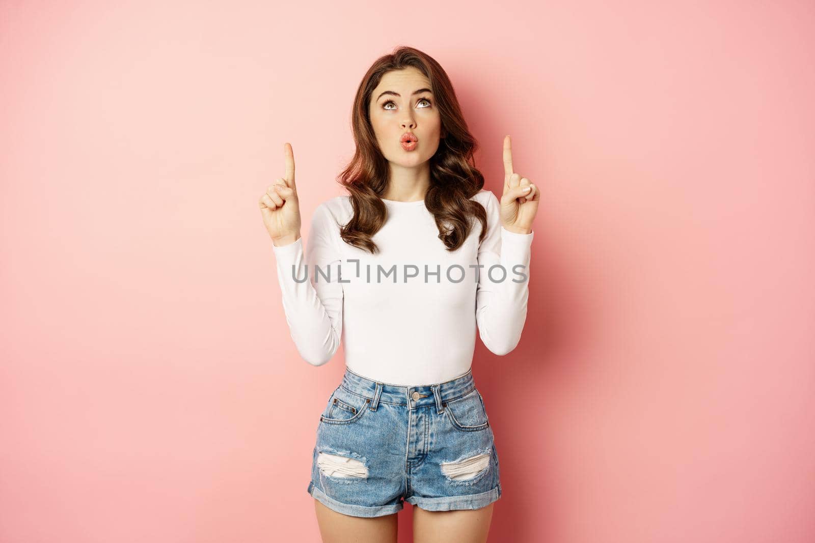 Portrait of surprised caucasian woman, stylish girl pointing fingers up and looking impressed, showing store logo, discount advertisement, pink background by Benzoix