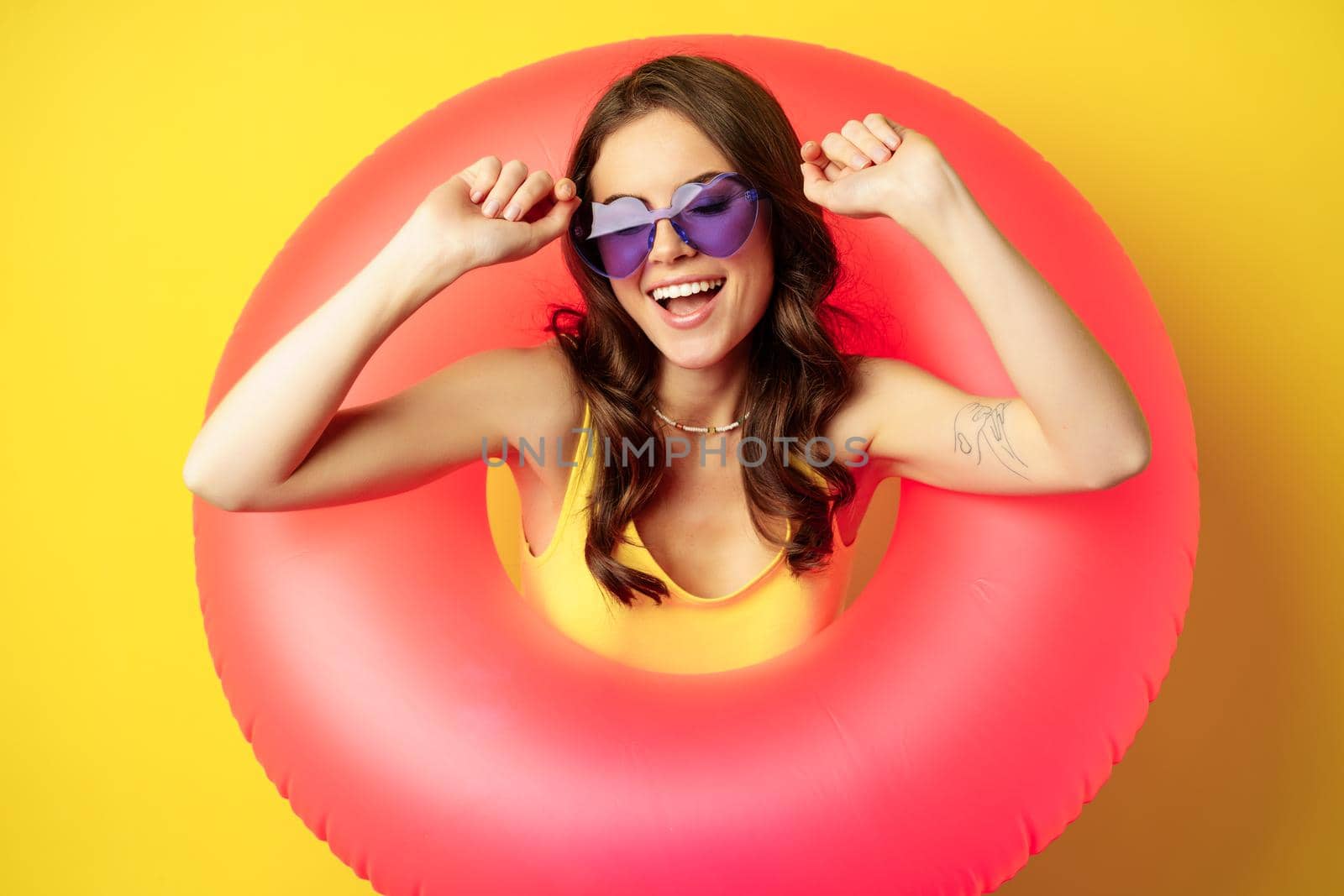 Close up portrait of stylish young woman, girl with swimming ring and summer sunglasses, ready for beach holiday, vacation, standing over yellow background by Benzoix