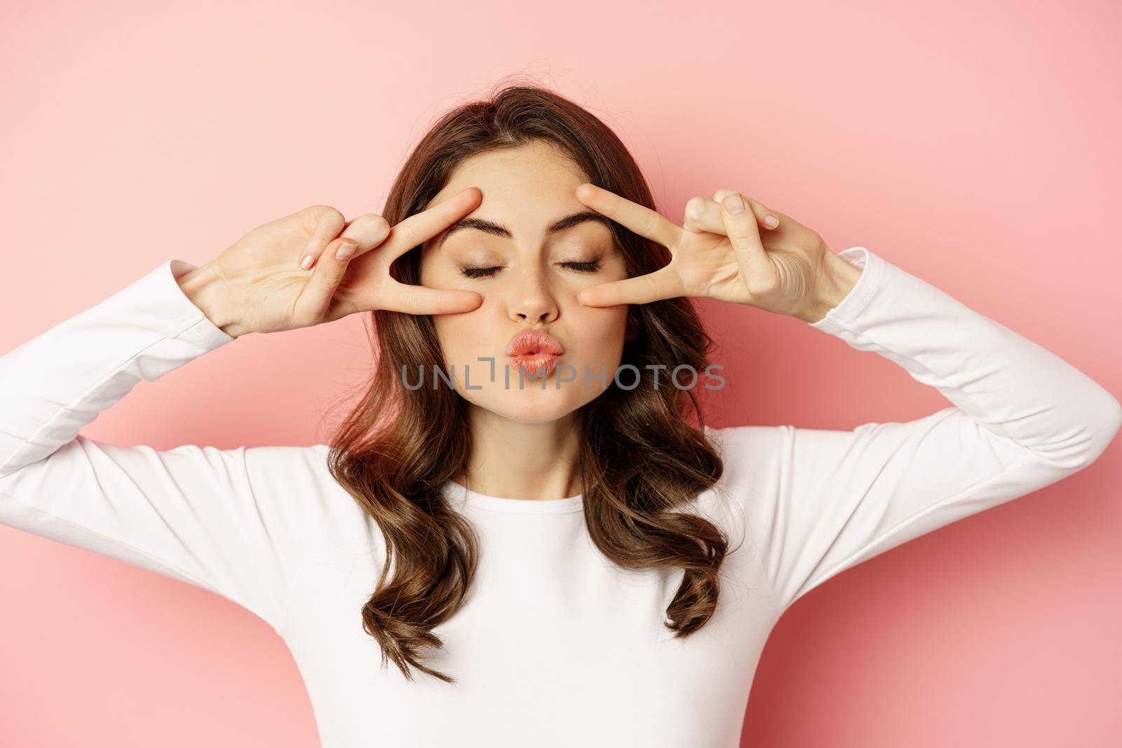 Close up face of young stylish girl, sassy woman showing happy, excited face expression, peace v-sign over eyes, standing against pink background by Benzoix