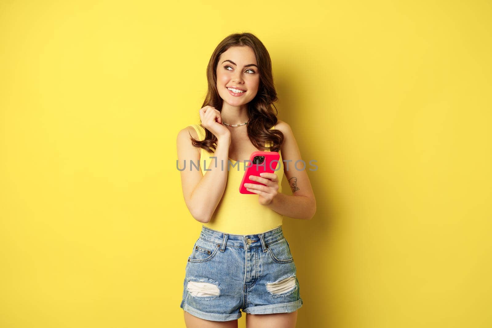 Technology and cellphone. Beautiful stylish girl order in online store, praying in application, using smartphone app and thinking, making decision, standing over yellow background by Benzoix