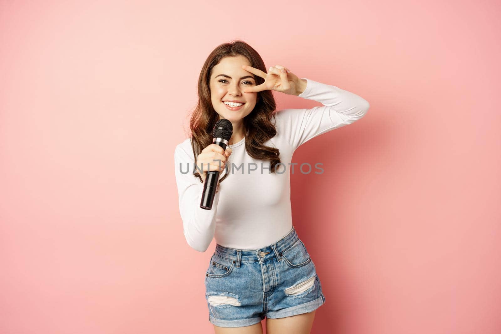 Party girl. Happy young woman singing in microphone, performing song, having fun at event, standing over pink background by Benzoix