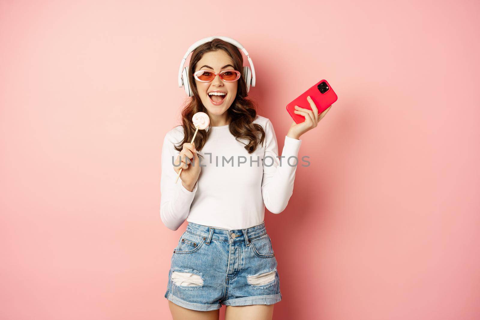 Summer girl laughing, eathing lolipop and listening music in headphones, dancing with smartphone against pink background by Benzoix