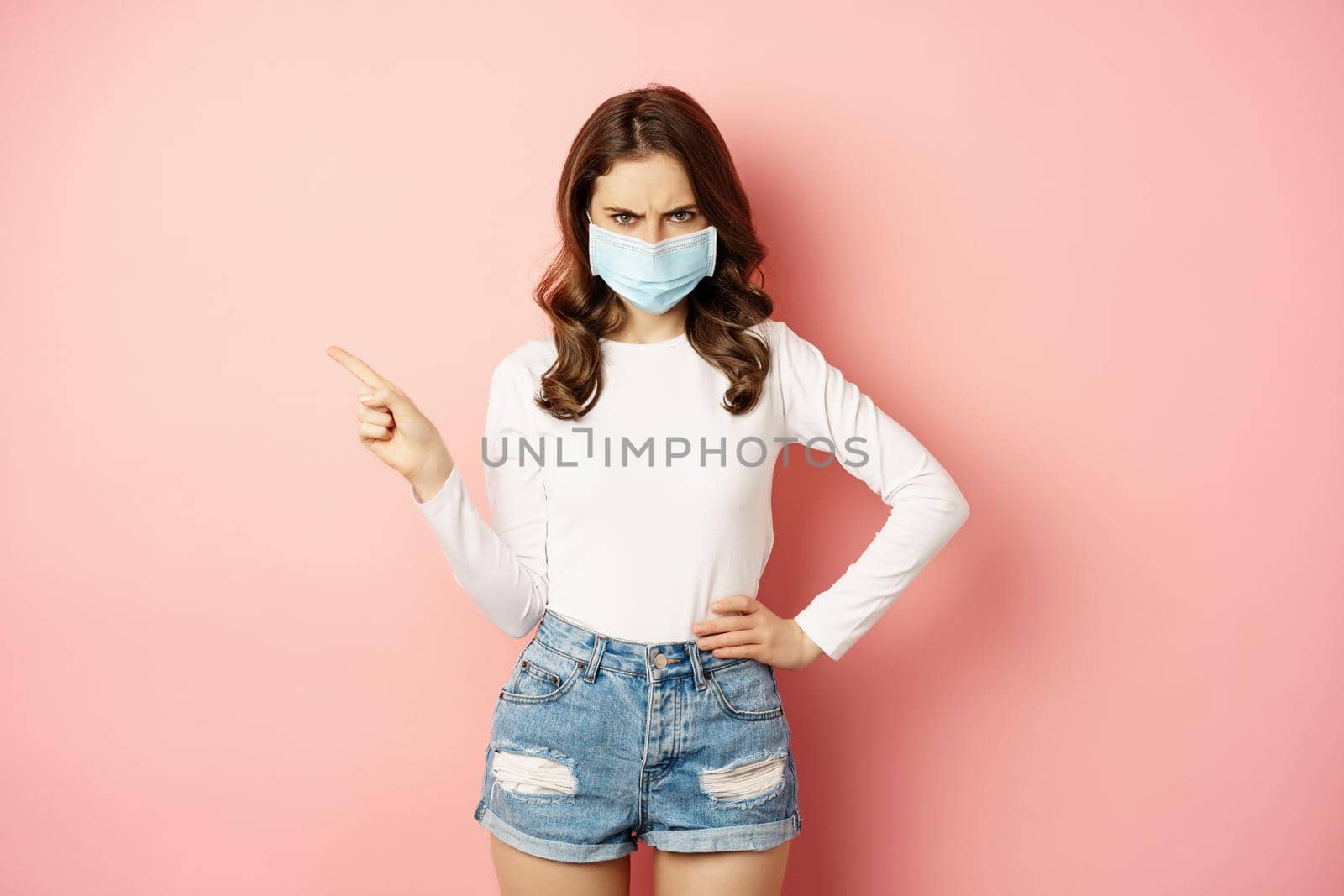 Angry young woman in medical face mask, frowning frustrated, pointing finger left, showing promo copy space, standing over pink background by Benzoix