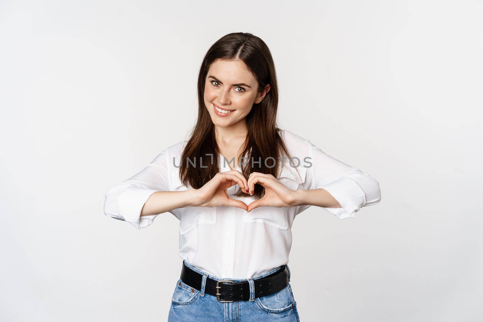 Lovely feminine woman showing heart sign, romantic gesture and smiling with care and tenderness, standing over white background.