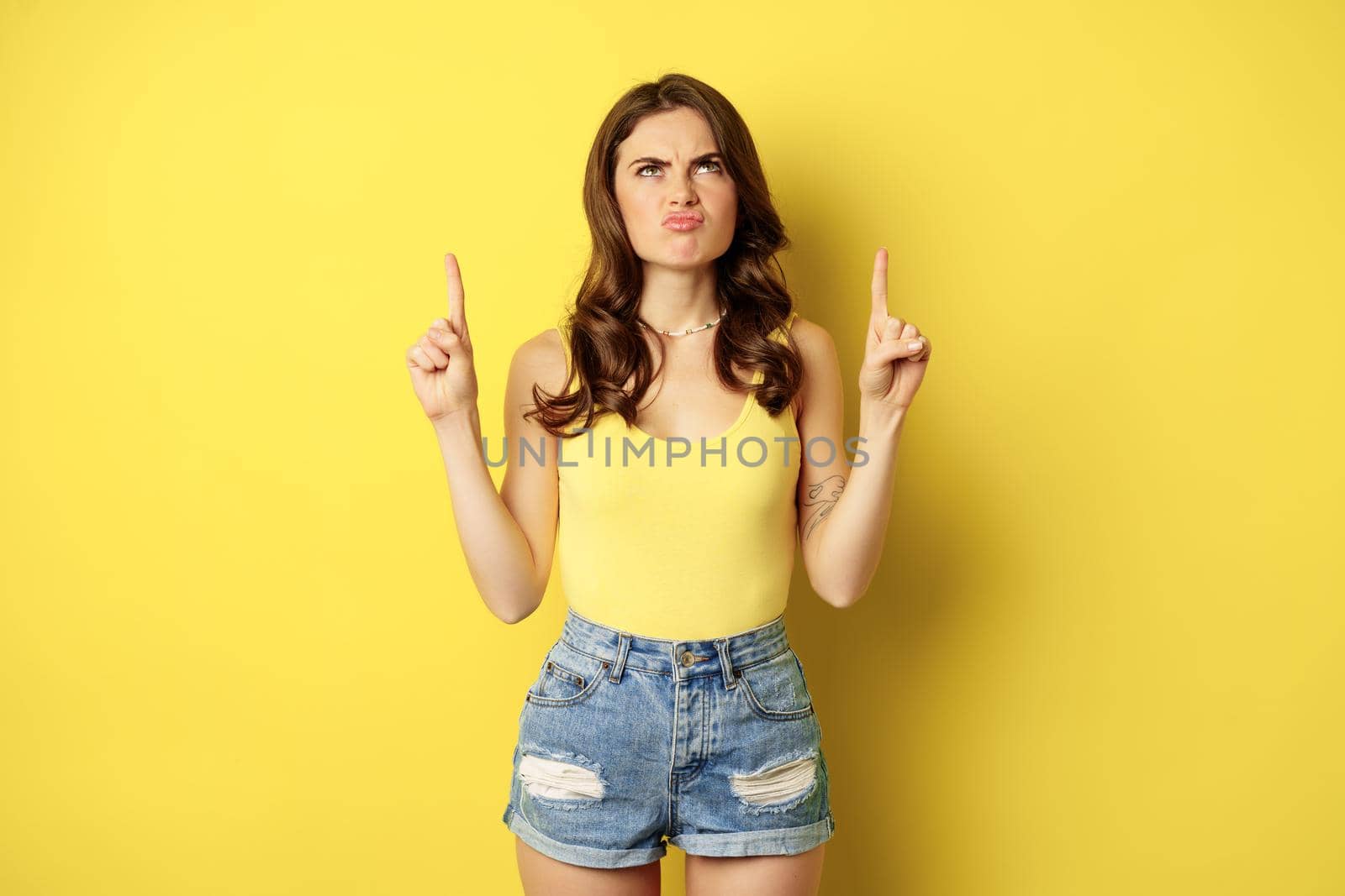 Disappointed girl grimacing, pointing fingers, showing logo, promo offer, banner or logo, standing over yellow background.