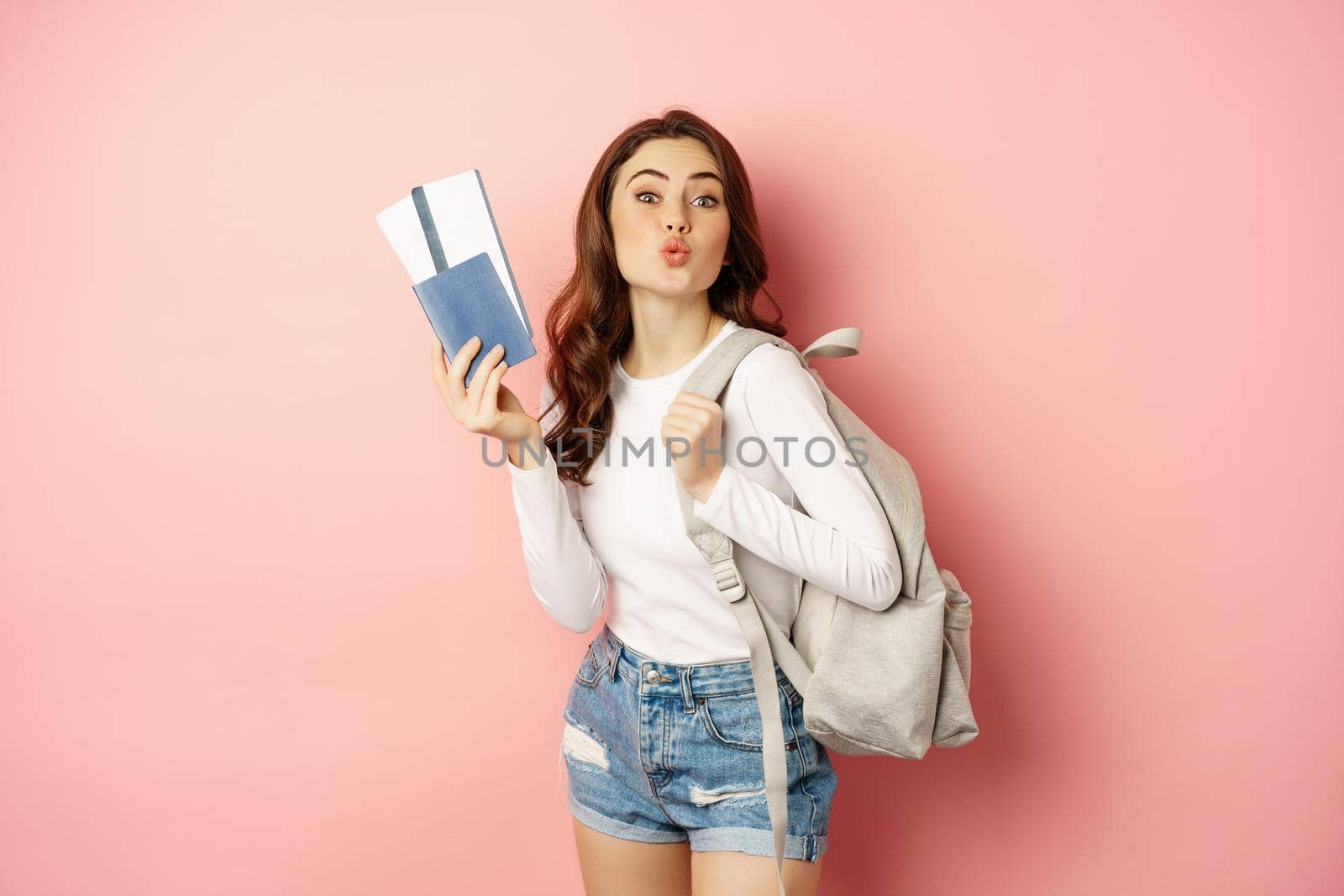 Beautiful girl on vacation, going on trip with passport and tickets, holding backpack. Young woman traveller backpacking, standing over pink background.