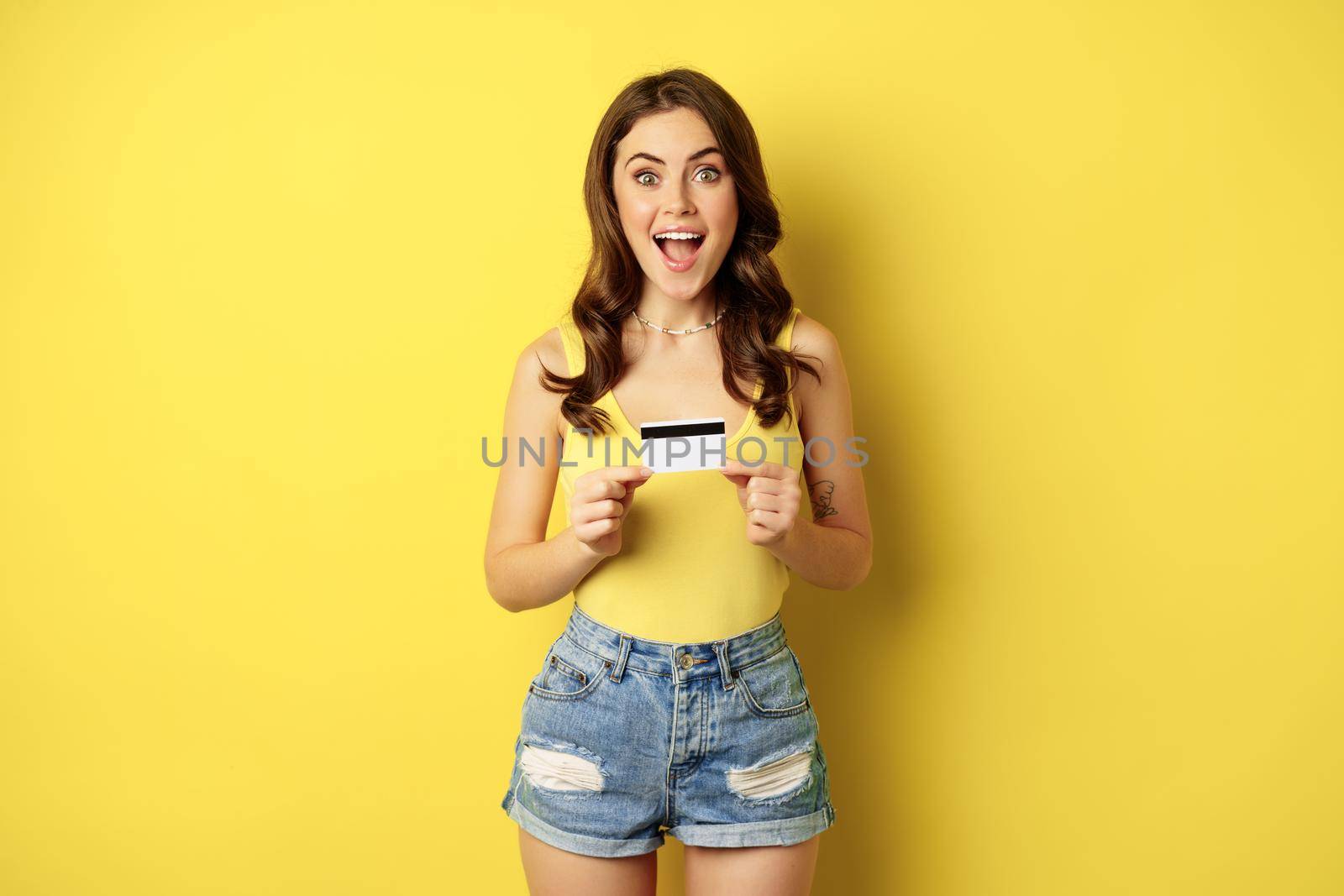 Enthusiastic young woman showing credit card, recommending bank, discount in store, contactless payment or cashback, standing over yellow background.