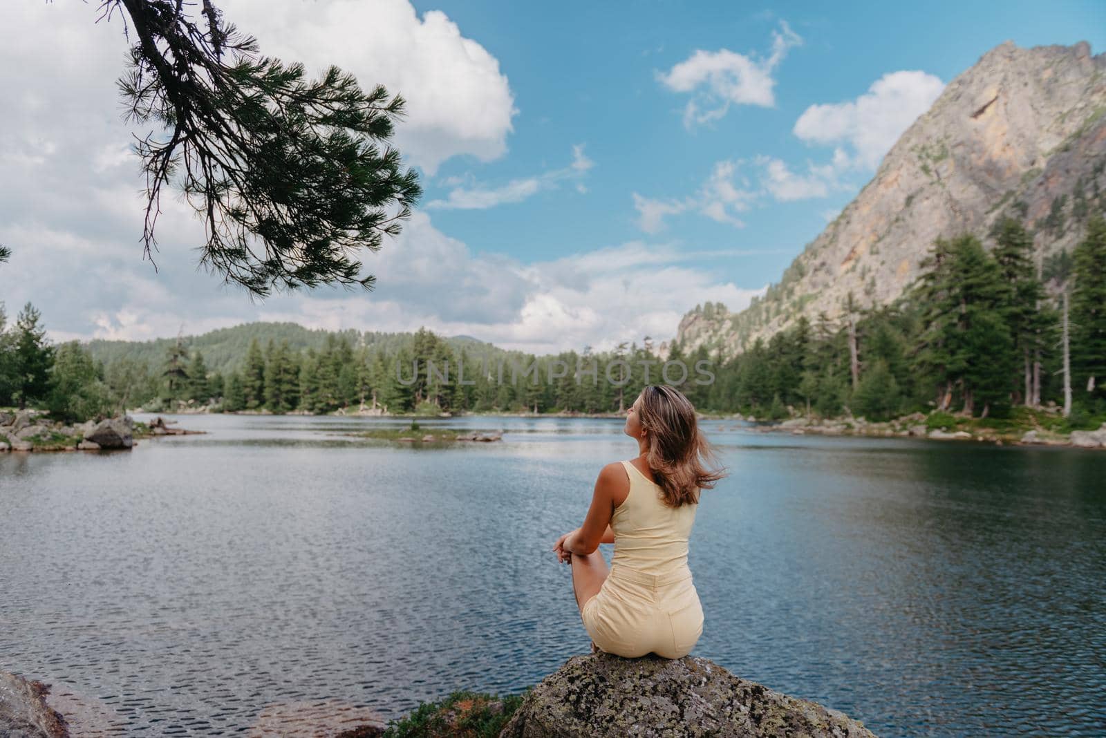 Tourist girl enjoys the magical view of the lake, coniferous forest and magical view sitting on big stone on the shore of a turquoise lake in the mountains. Hiking in the Natural Park. Cute girl tourist sitting on a large stone by the lake. The water is clear, stones are visible under the water. by Andrii_Ko