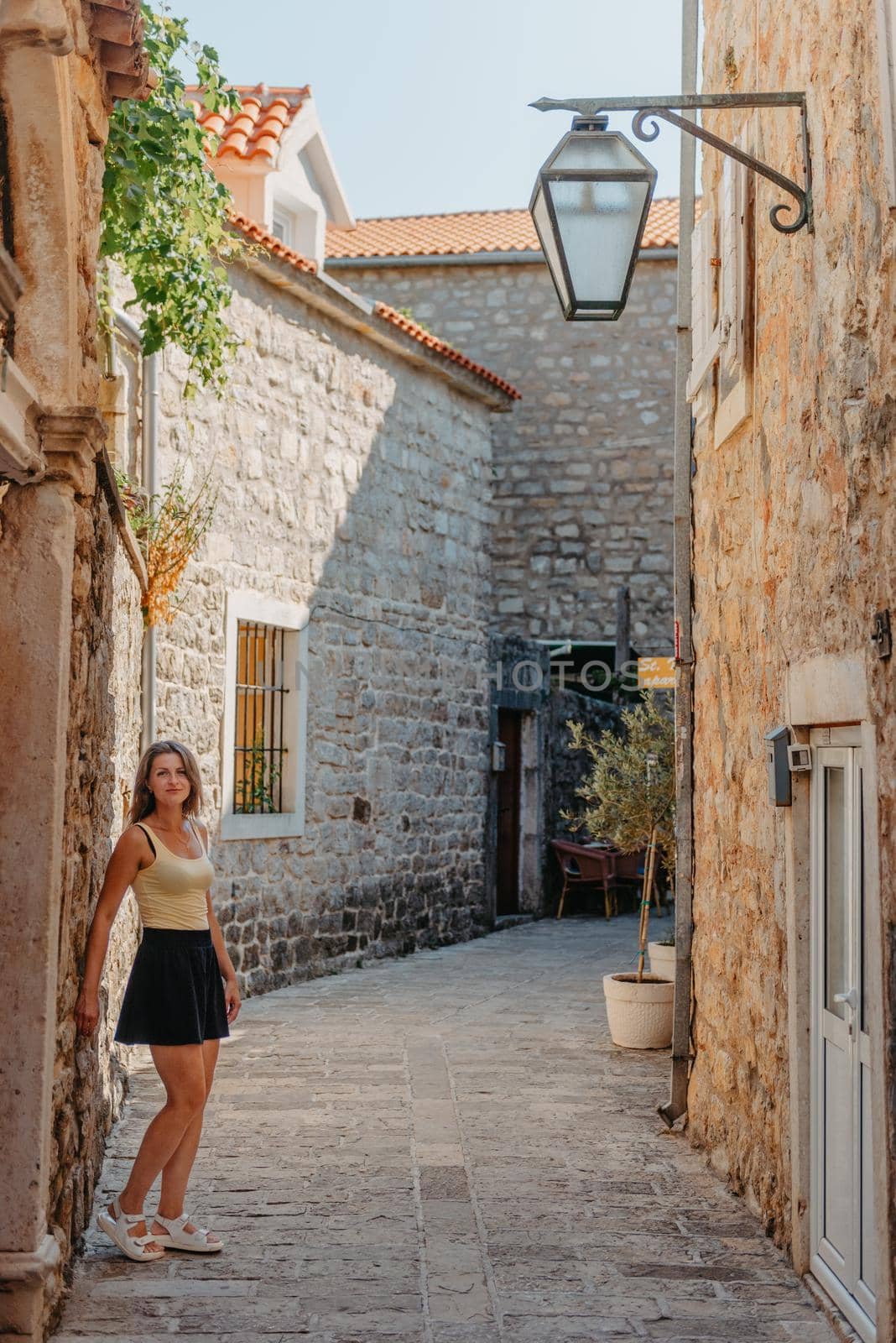 Girl Tourist Walking Through Ancient Narrow Street On A Beautiful Summer Day In MEDITERRANEAN MEDIEVAL CITY, OLD TOWN BUDVA, MONTENEGRO. Young Beautiful Cheerful Woman Walking On Old Street At Tropical Town. Pretty Girl Looking At You And Smiling by Andrii_Ko