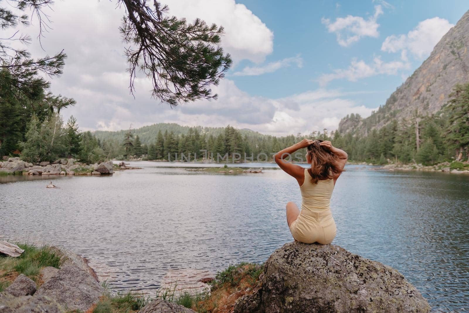 Tourist girl enjoys the magical view of the lake, coniferous forest and magical view sitting on big stone on the shore of a turquoise lake in the mountains. Hiking in the Natural Park. Cute girl tourist sitting on a large stone by the lake. The water is clear, stones are visible under the water. by Andrii_Ko