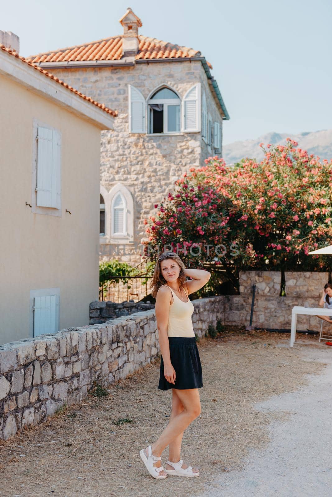 Girl Tourist Walking Through Ancient Narrow Street On A Beautiful Summer Day In MEDITERRANEAN MEDIEVAL CITY, OLD TOWN BUDVA, MONTENEGRO. Young Beautiful Cheerful Woman Walking On Old Street At Tropical Town. Pretty Girl Looking At You And Smiling by Andrii_Ko