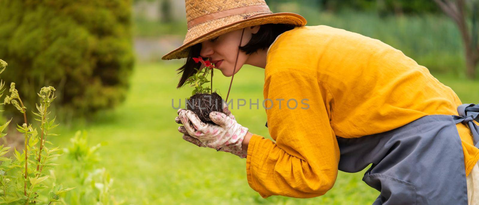 A young woman gardener in a straw hat and hands in gloves is holding a petunia flower in a peat pot, sniffing the flower and smiling before planting a seedling