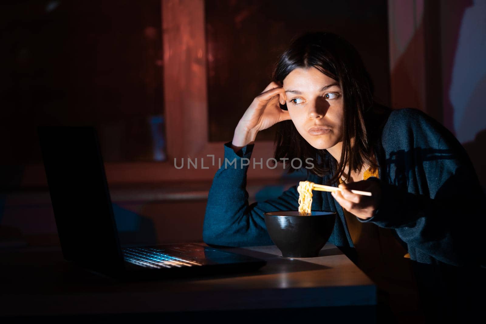 Young girl works at night at home with laptop and eats noodles, asian food with chopsticks. A woman watches a show, a movie and has dinner with ordered food from a cafe.