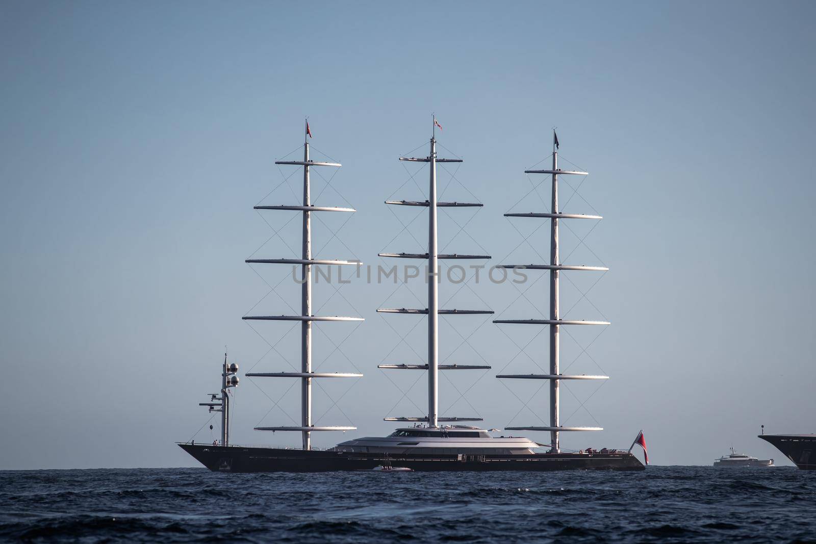 A huge sail yacht of conceptual design with three masts stands in the bay near Monaco at sunny day, the sails are lowered, the yacht spins around its anchor, left side of the sailboat, clear weather. High quality photo