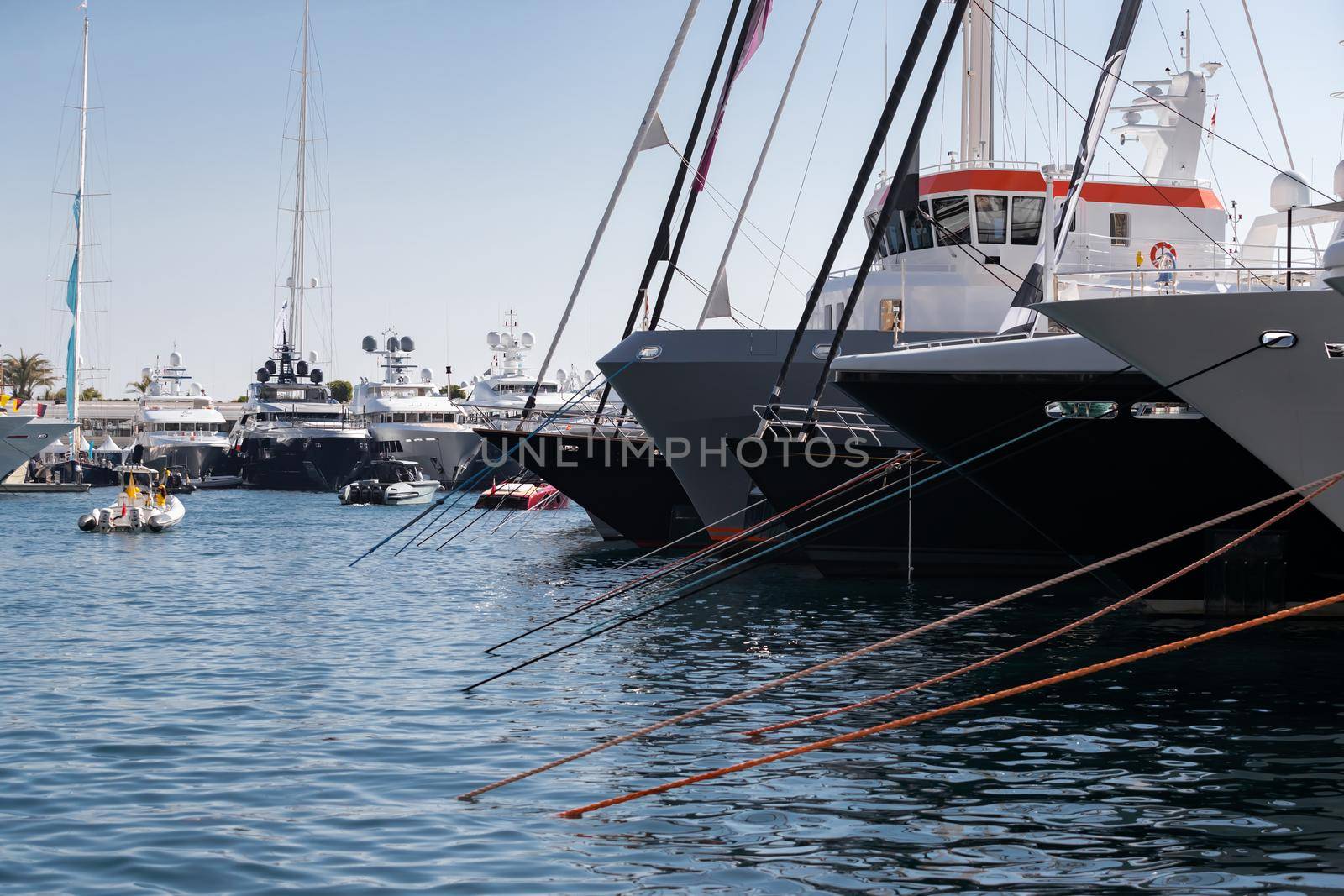 A lot of huge yachts are in port of Monaco at sunny day, Monte Carlo, mountain is on background, big masts of sail yacht, motorboat and mega yachts are moored in marina, sun reflection on glossy board by vladimirdrozdin