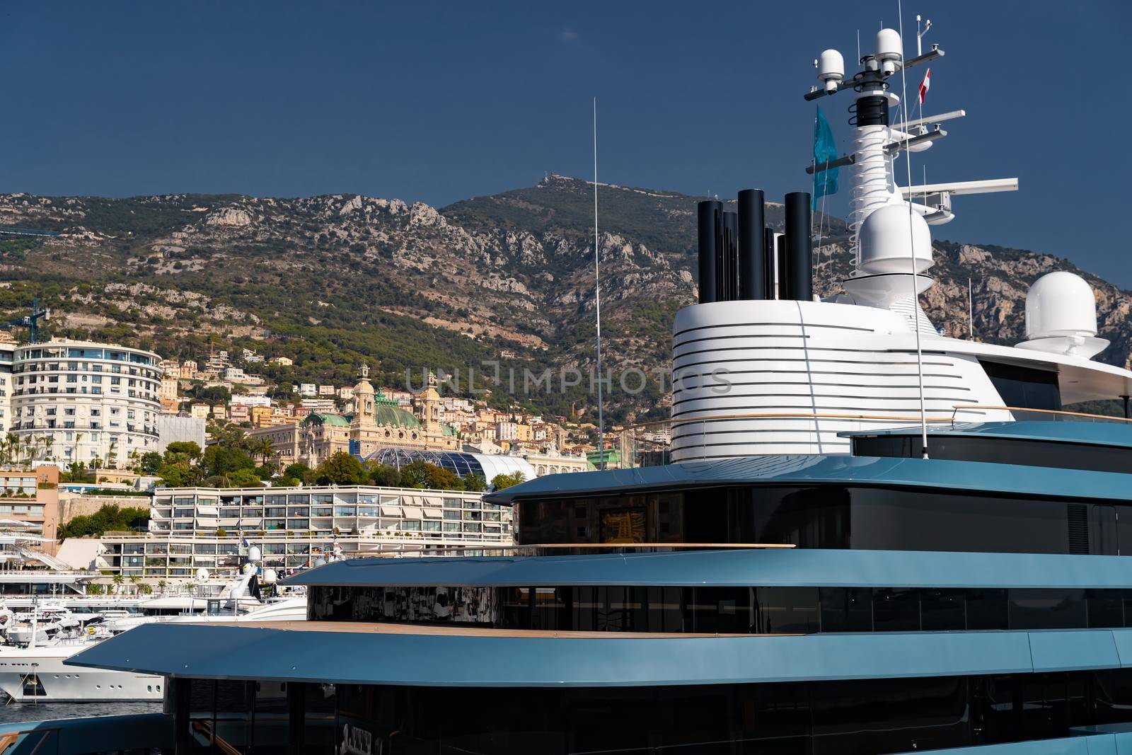 Landmarks of Monaco through decks of huge yacht of blue color in port Hercule at sunny day, glossy board of the motor boat, sun reflection on glossy board, mountains are on background. High quality photo