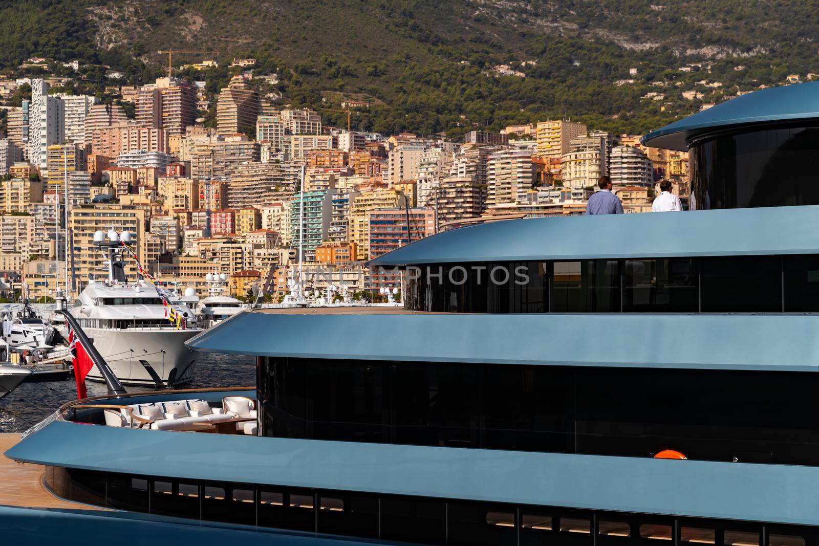 Landmarks of Monaco through decks of huge yacht of blue color in port Hercule at sunny day, glossy board of the motor boat, sun reflection on glossy board, mountains are on background. High quality photo