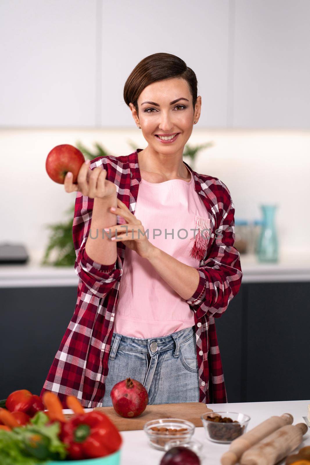 Apple in hand happy woman select fruits cooking at new kitchen looking at camera. Housewife cooking apple pie standing at the kitchen wearing plaid shirt with a short hair.