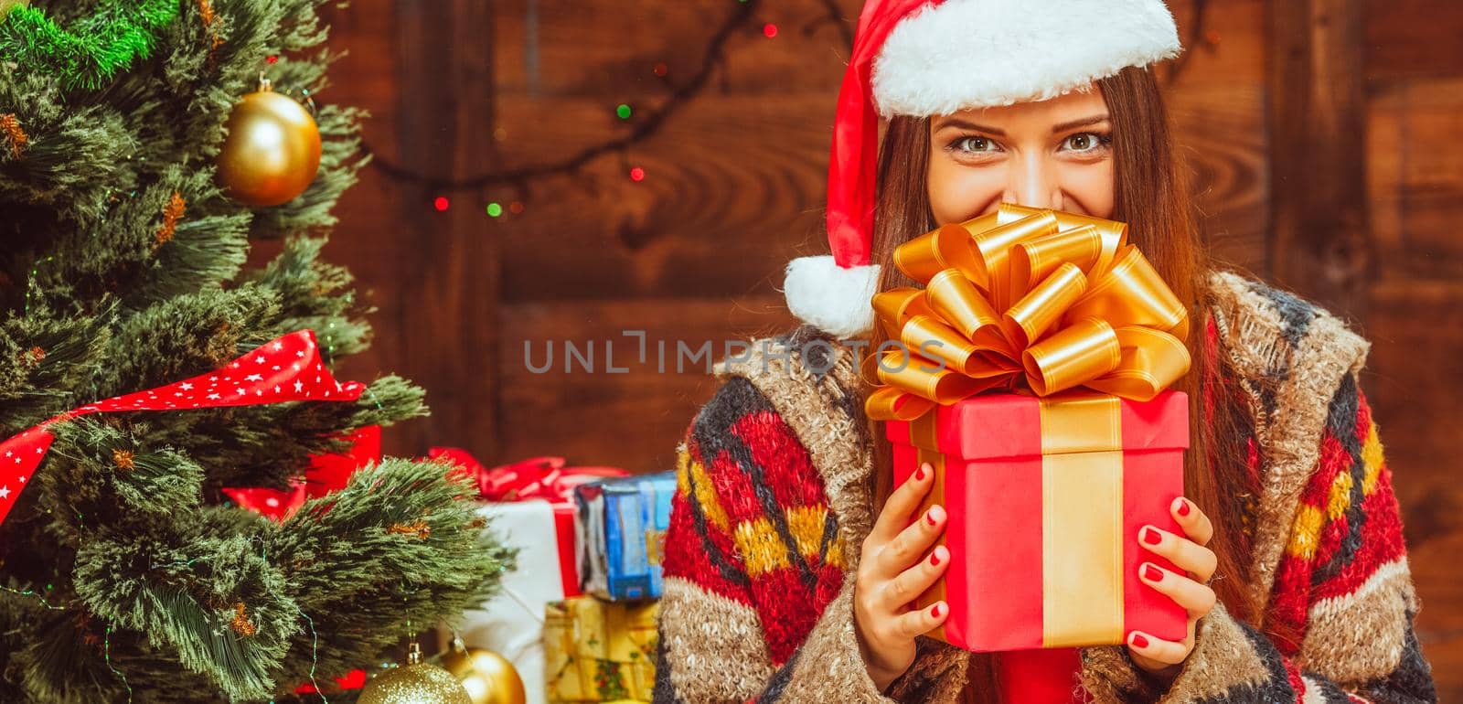 Christmas Lady has a Red Square Gift Box with a Gold Ribbon on its Sides and a Large Loopy Bow on its Top. Young Woman Holds a Christmas Present in her Hands and Covers the Lower Part of her Face. Copy Space. Christmas Tree Background. Wood Wall. High quality photo