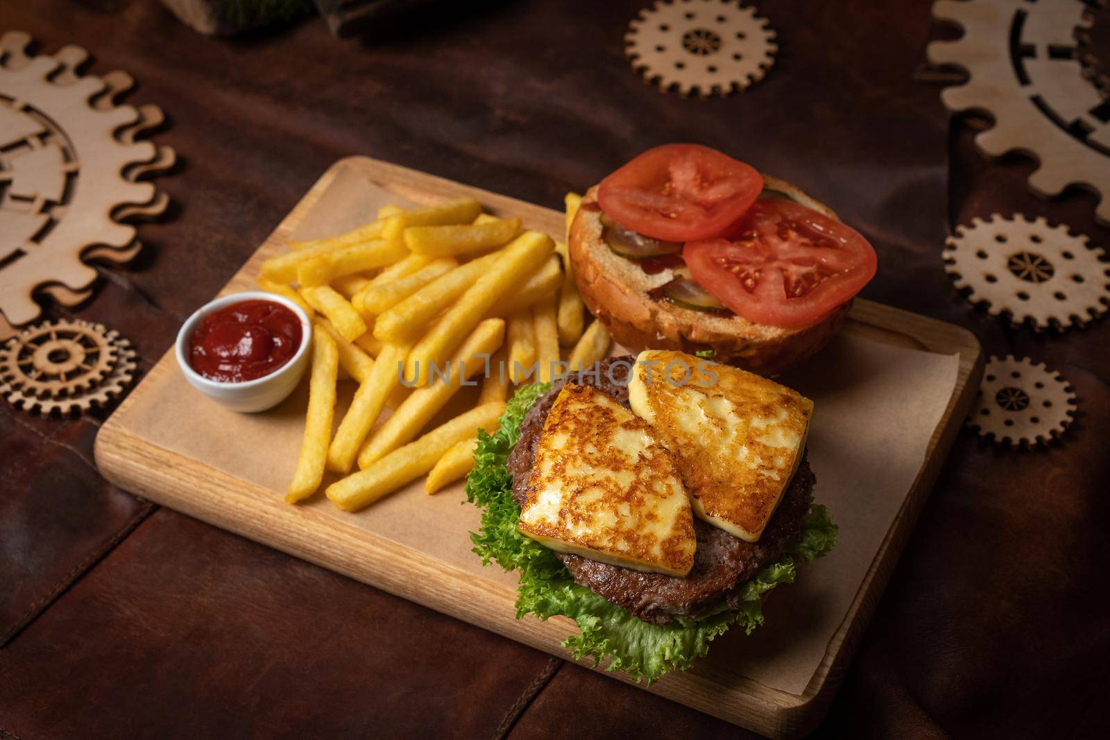 Disassembled beef burger with fried cheese, slices tomatoes served with french fries on a decorative wooden board with tomato sauce with wooden decorative pieces of simple mechanism.