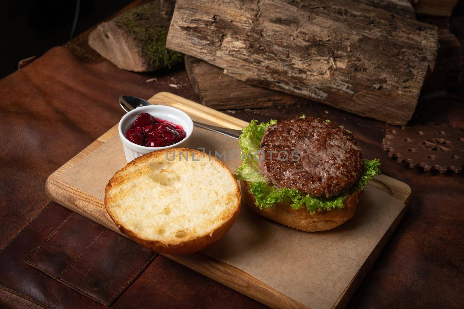 Fresh craft beef burger with cherry sauce in a sauce bowl, fresh lettuce with lead bun laying next with wooden lodges as decoration. Restaurant concept. Street food concept.