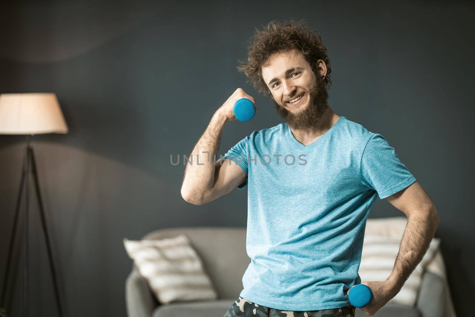 Funny handsome man use dumbbell to exercise at home laughing looking at camera. Funny disheveled hair man with a mess beard doing exercise at home. Concept of healthy life.