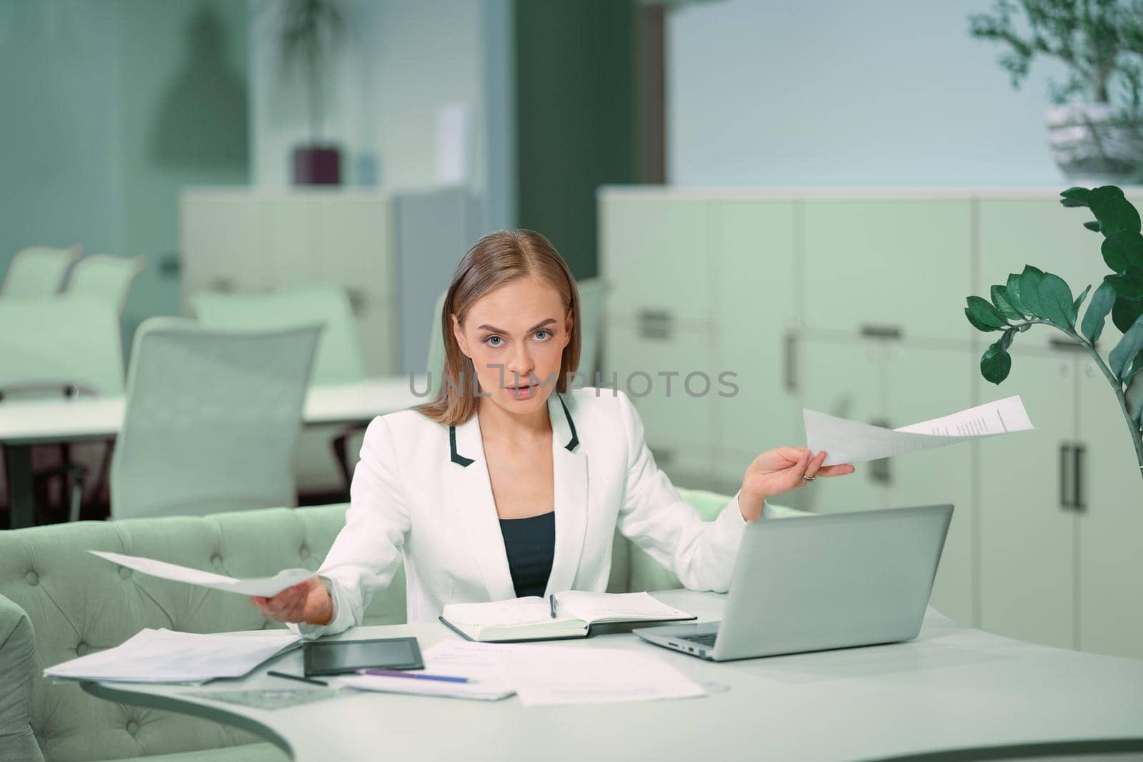 Dissatisfied or tired blond businesswoman working with documents sitting in front of laptop wearing white official suit. Office worker looking at camera sitting at her working place.