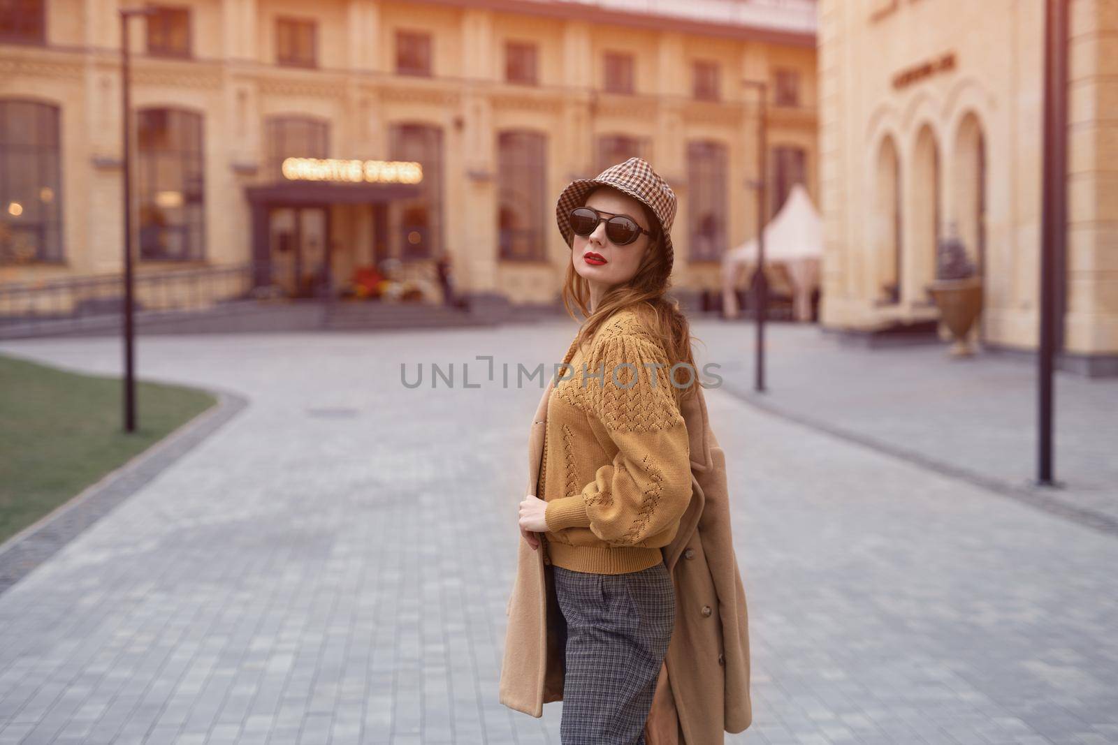 Turned from behind fashion model young woman in sunglasses and hanging on a shoulder autumn beige coat walking on the street happily posing for the camera. Tinted photo.