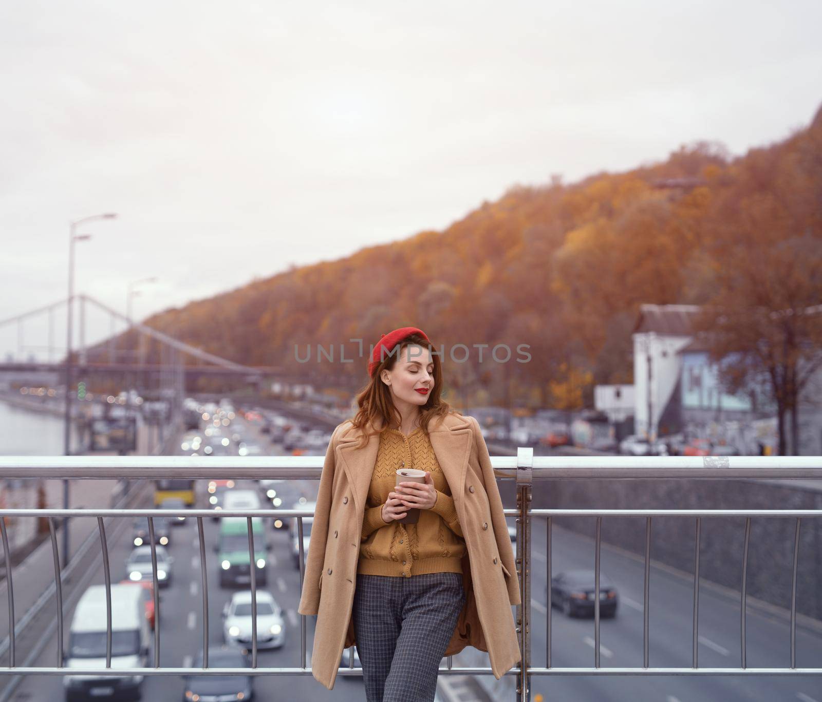 Charming Parisian young woman drinks coffee using reusable mug looking at camera smiling standing at pedestrian bridge with cars on the road. Looking happy fashion girl in a red beret and beige coat.