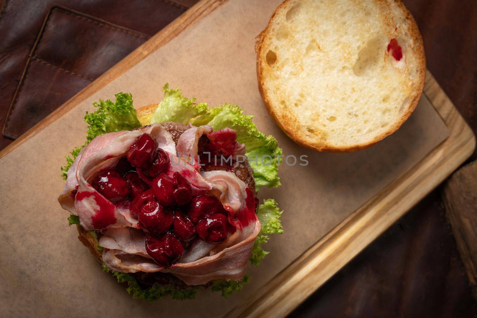 Fresh craft beef burger with fried slices of bacon and cherry sauce on top, fresh lettuce with lead bun laying next to it. Restaurant concept. Street food concept.