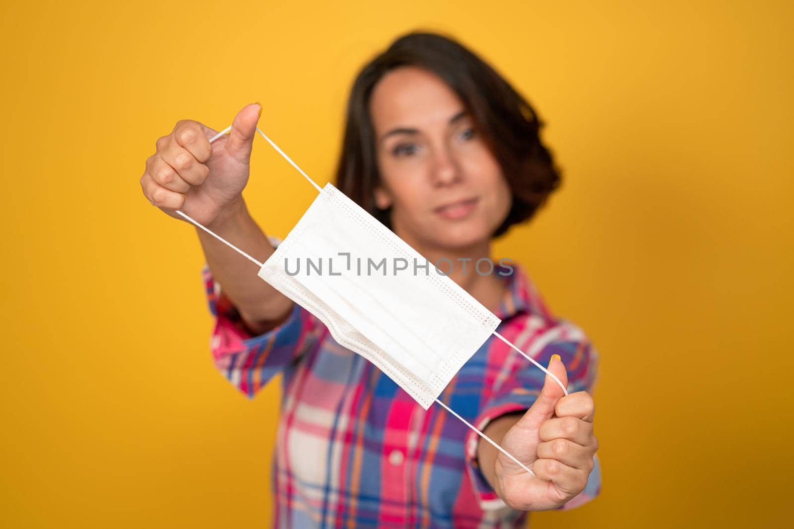 Cute young woman holds protective mask in foreground. Isolated on yellow background. Quarantine concept. Copy space.