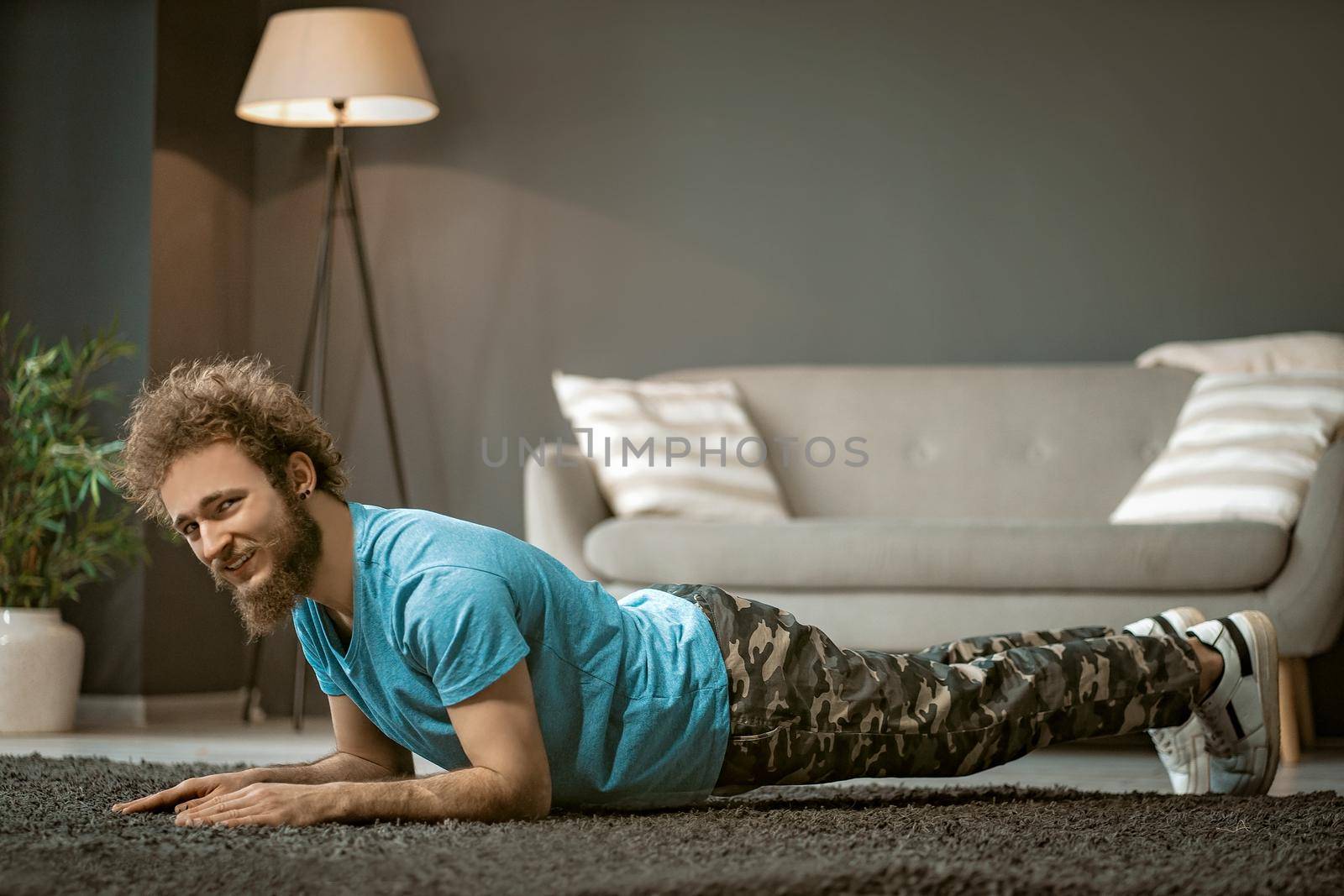 Cheerful Guy in a Blue T-shirt Does Plank Indoor. Young Man Does Up-downs to Get Fit. Medium close-up. High quality photo