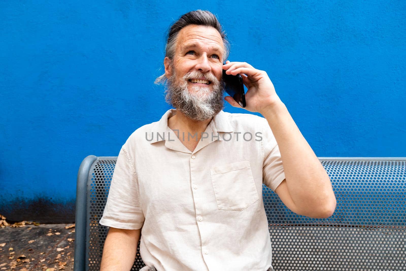 Mature caucasian man with beard sitting on a bench using mobile phone outdoors. Blue background by Hoverstock