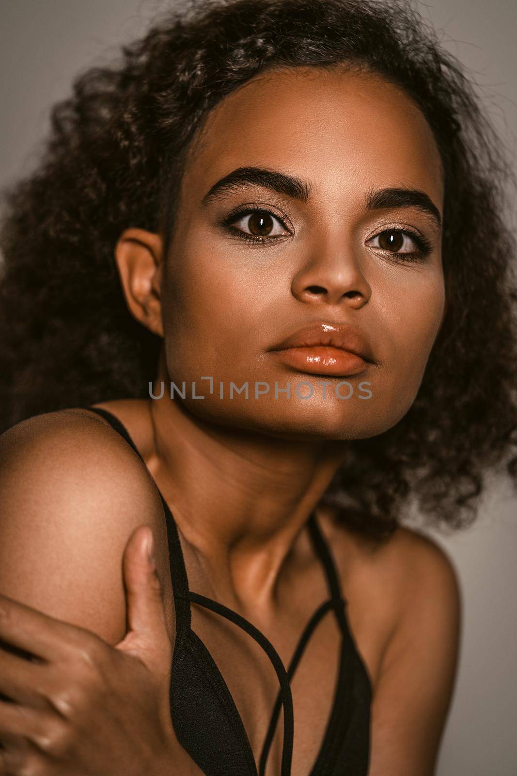 Gentle passionate African American young woman standing covered with crossed hands in black bare shoulder top isolated on grey background. Human emotions, facial expression concept.