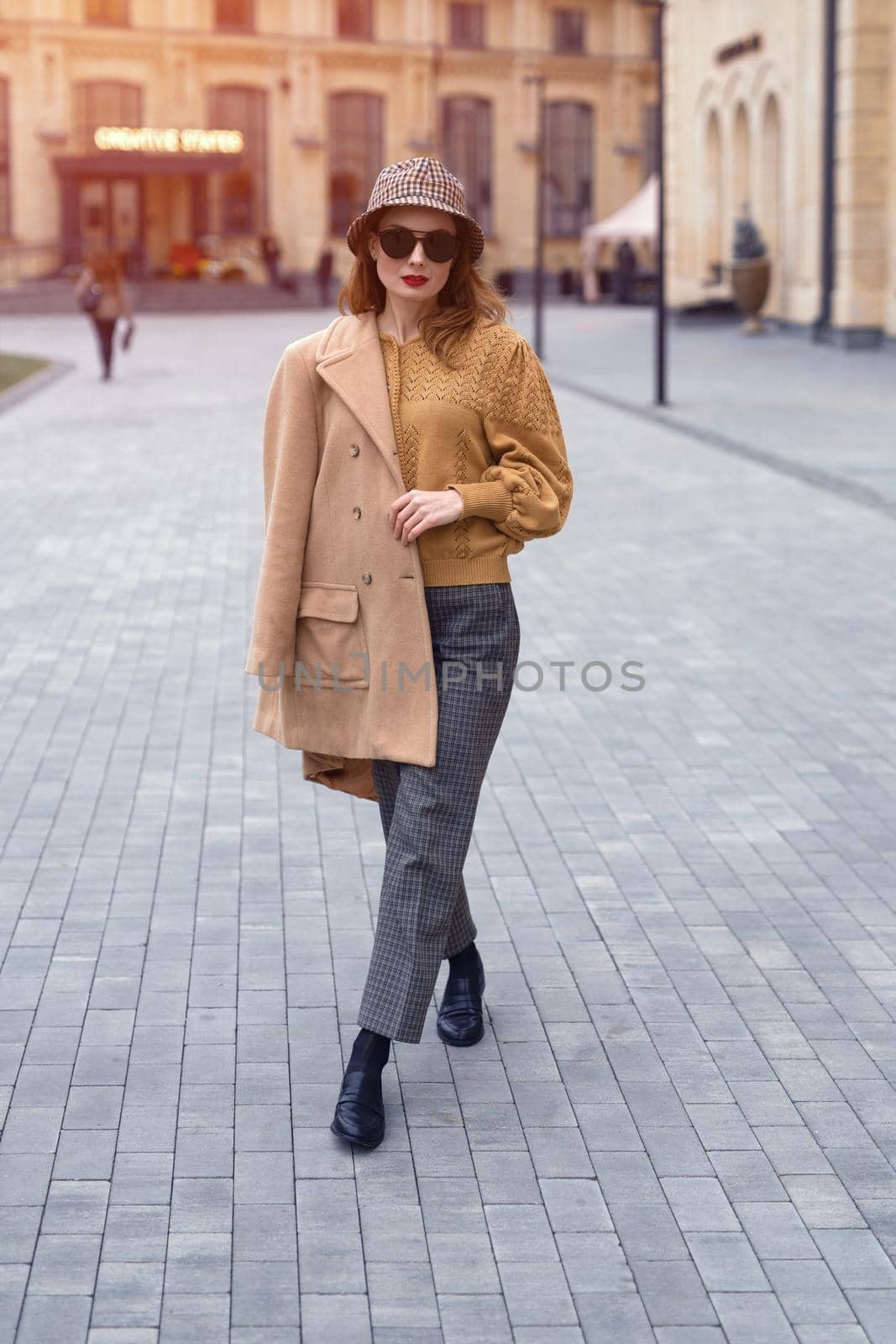 Outdoor portrait of charming spring time young french woman wearing sunglasses walking on camera with old town on background. Stylish female tourist standing at the city center square. Toned image.