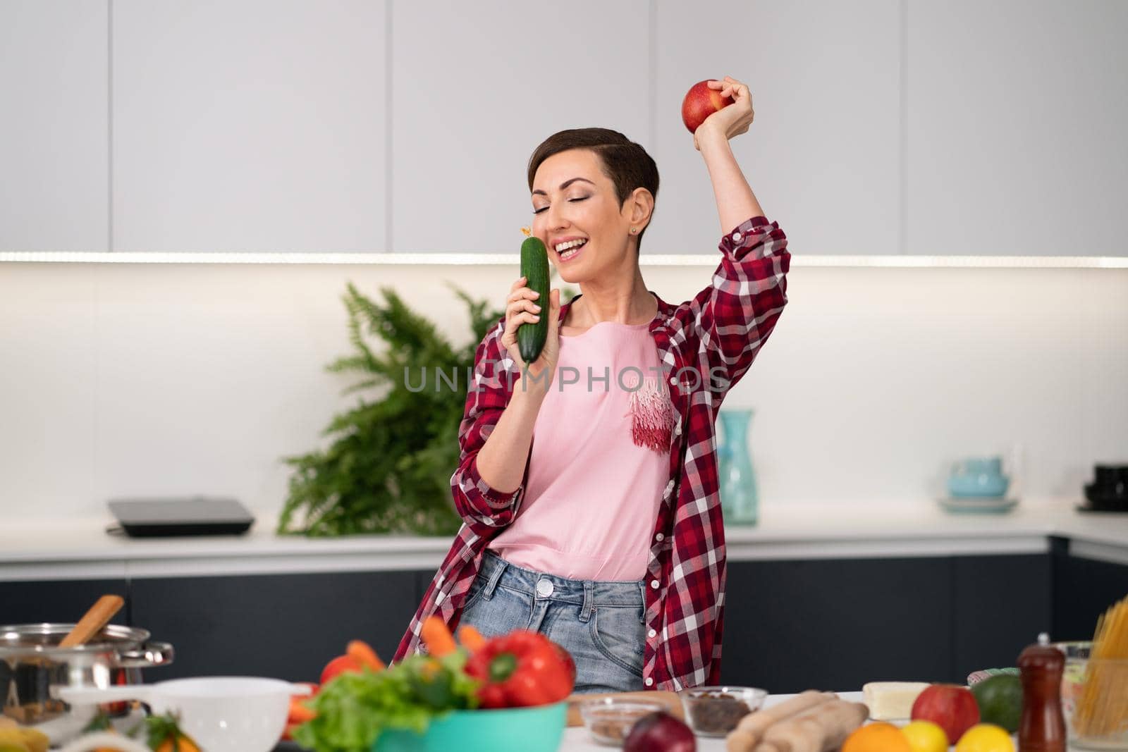Young pretty housewife with a bob hairstyle cooking dinner or lunch standing in the kitchen choosing fresh vegetables smelling cucumber and apple in hand. Healthy food at home.