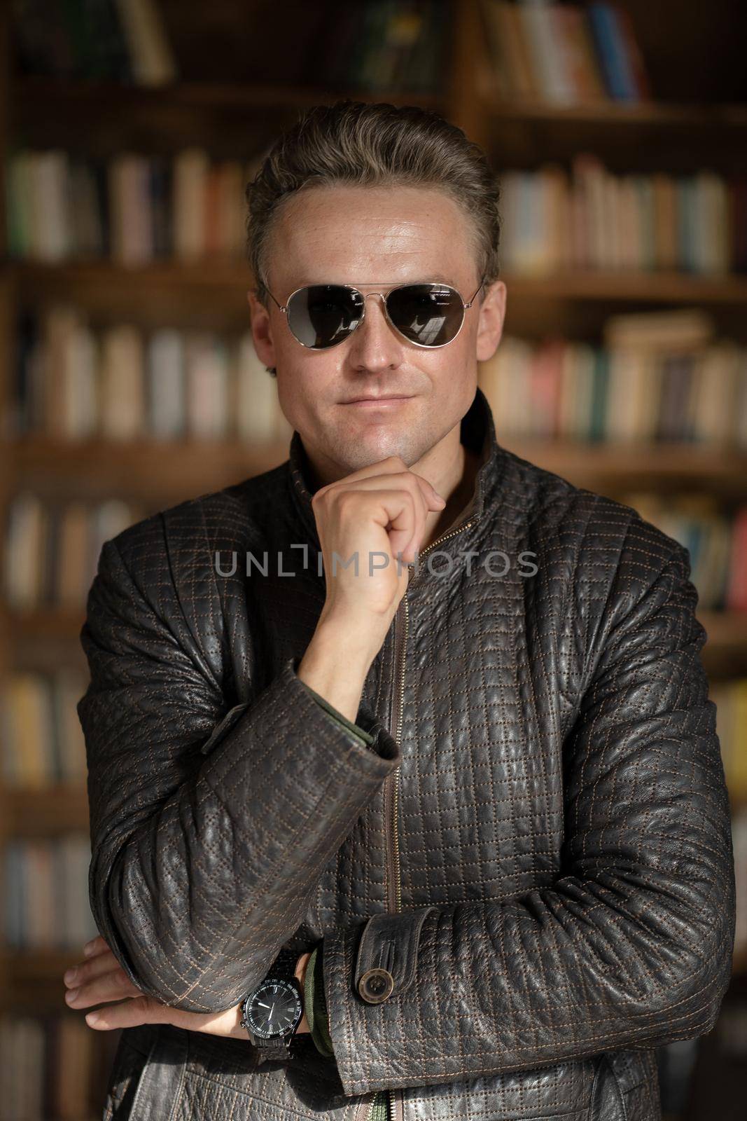 Brutal Fair-haired Guy in Modern Sunglasses Posing in his Business Office .White Man in Lather Jacket. Close-up Portrait. Book Shelves Background. High quality photo