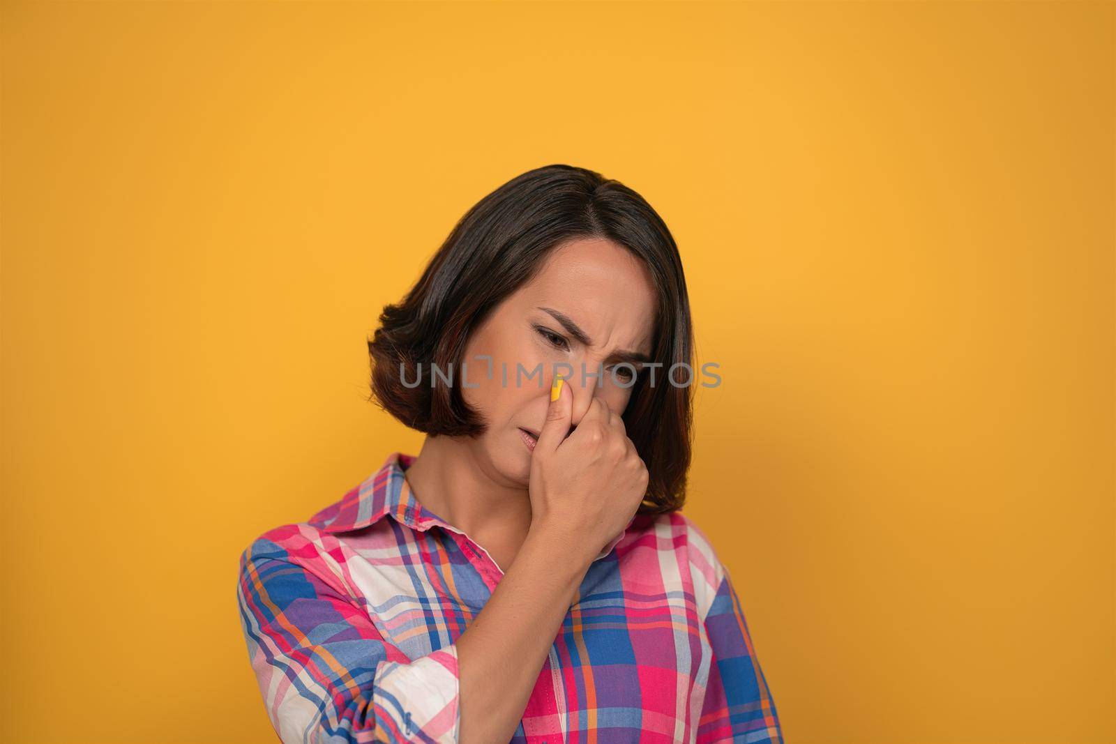 Bad smelling gesture closed nose cute looking woman shows her to her interlocutor, wearing plaid shirt. Isolated on yellow background.