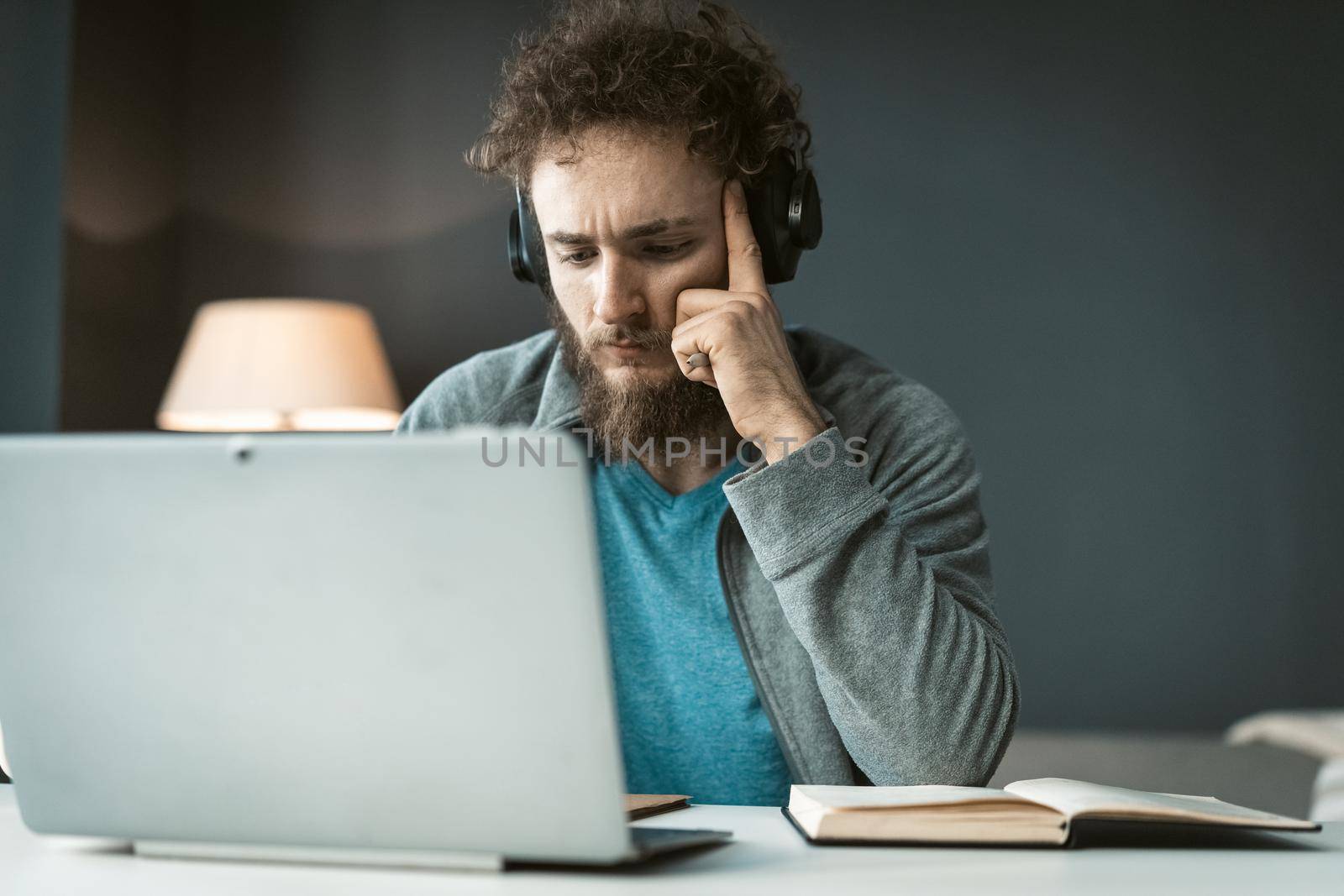 Serious young freelancer man working at laptop sitting at the desk at home office and taking notes. Remote working pensive young man working at home. Work from home concept.