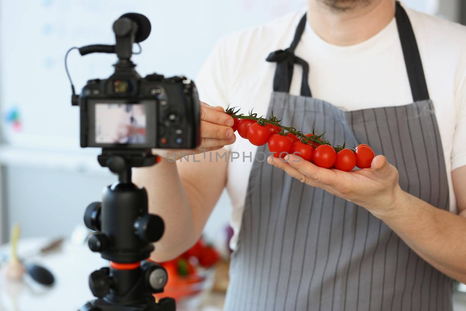 Close-up of vlogger chef showing ripe tomatoes ingredient, man in apron recording red vegetable on camera for culinary vlog. Hobby, content, cook concept
