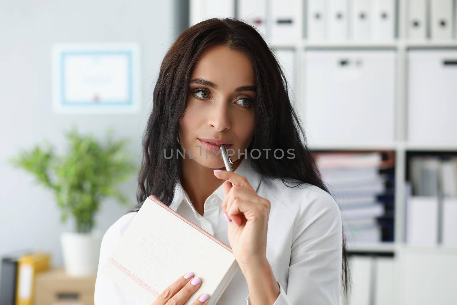 Portrait of woman holding silver pen on mouth, thinking about new ideas to journal. Female in suit at workspace with organizer diary. Creativity concept