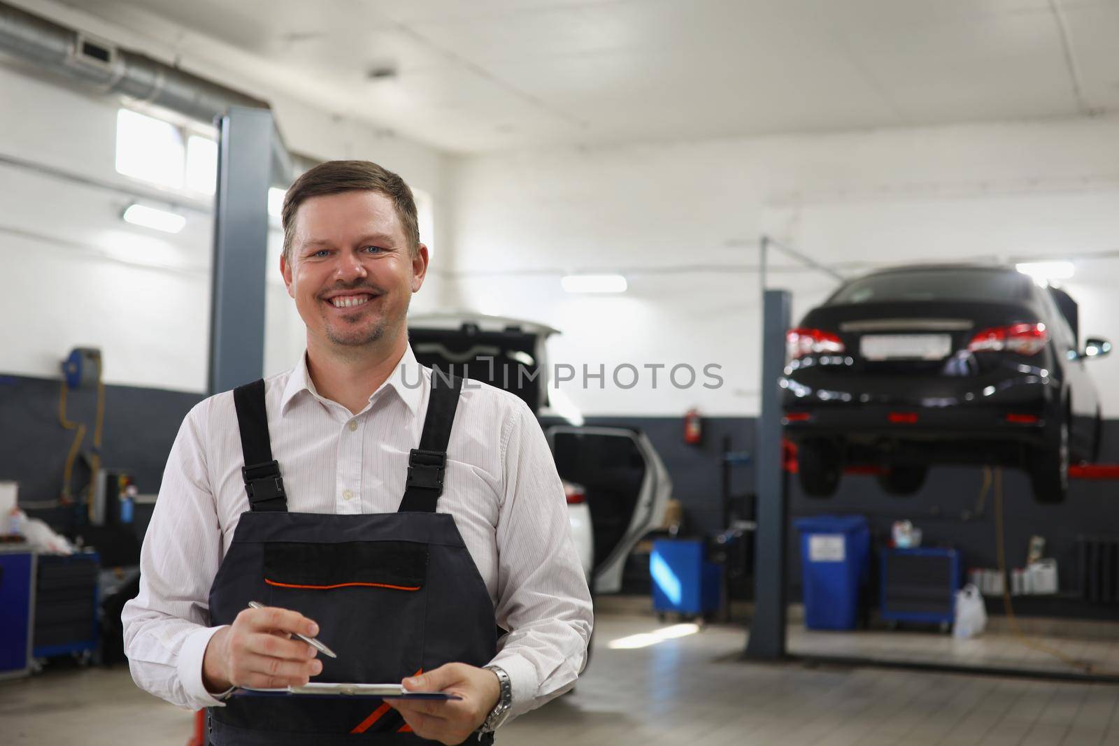 Smiling qualified maintenance center worker in uniform, man posing on cars background at work by kuprevich