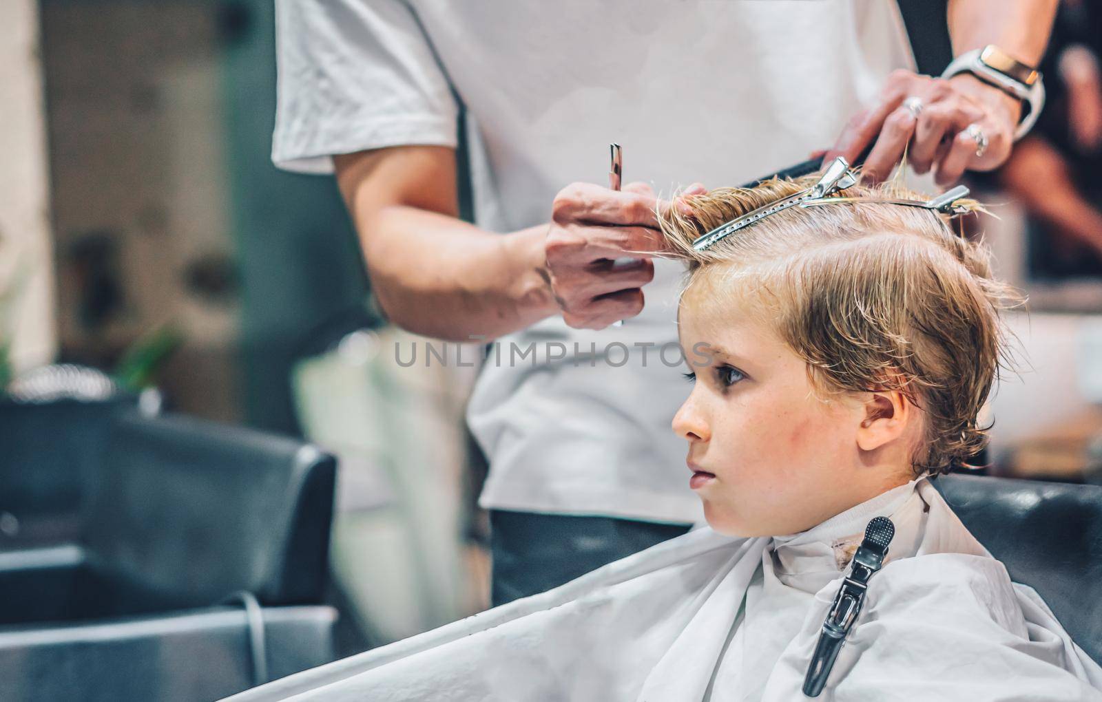 Lifestyle photo Barber shop blond boy customer. Hairdresser man work move hand comb tool scissors dryer clip white cape. Modern style kid short hair cut. Fashion business service for children.