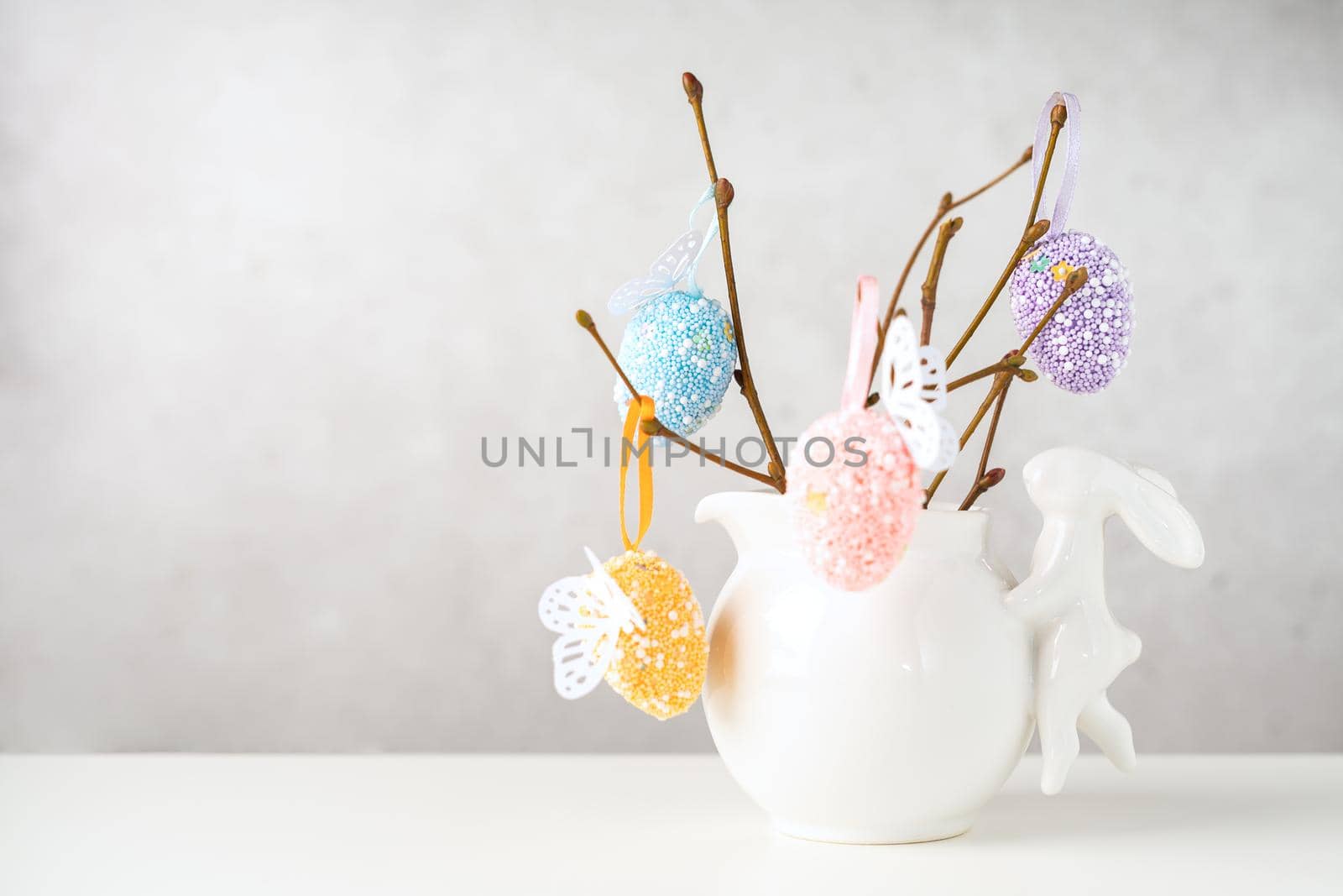 Home interior with easter decor. Vase with willow tree branches with Easter eggs and bunny on white table and background with copy space.