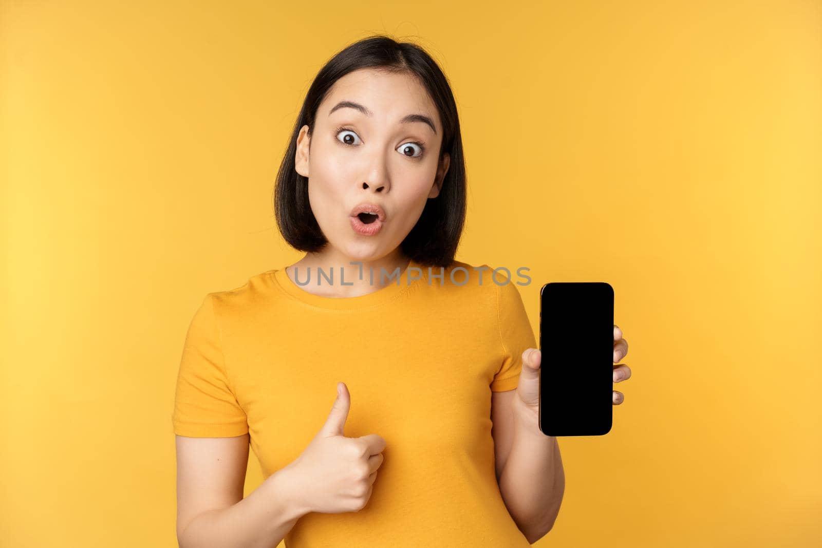 Excited asian woman showing mobile phone screen, thumbs up, like smth good, recommending smartphone application, standing over yellow background by Benzoix