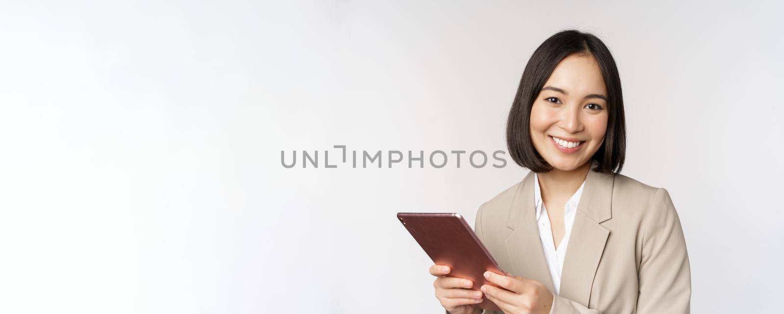 Image of asian businesswoman, saleswoman holding digital tablet and smiling, working with gadget, standing in suit over white background by Benzoix