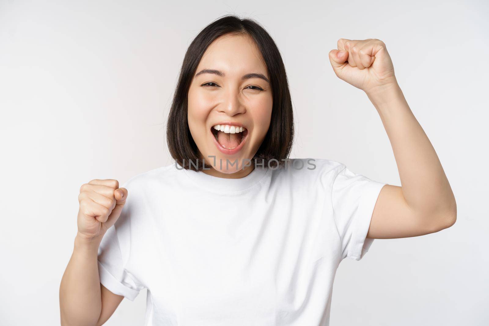 Portrait of enthusiastic asian woman winning, celebrating and triumphing, raising hands up, achieve goal or success, standing over white background by Benzoix