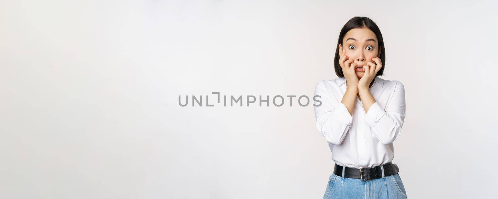 Portrait of scared asian woman watching smth scary, biting fingers on hands and looking at camera frightened, standing over white background by Benzoix