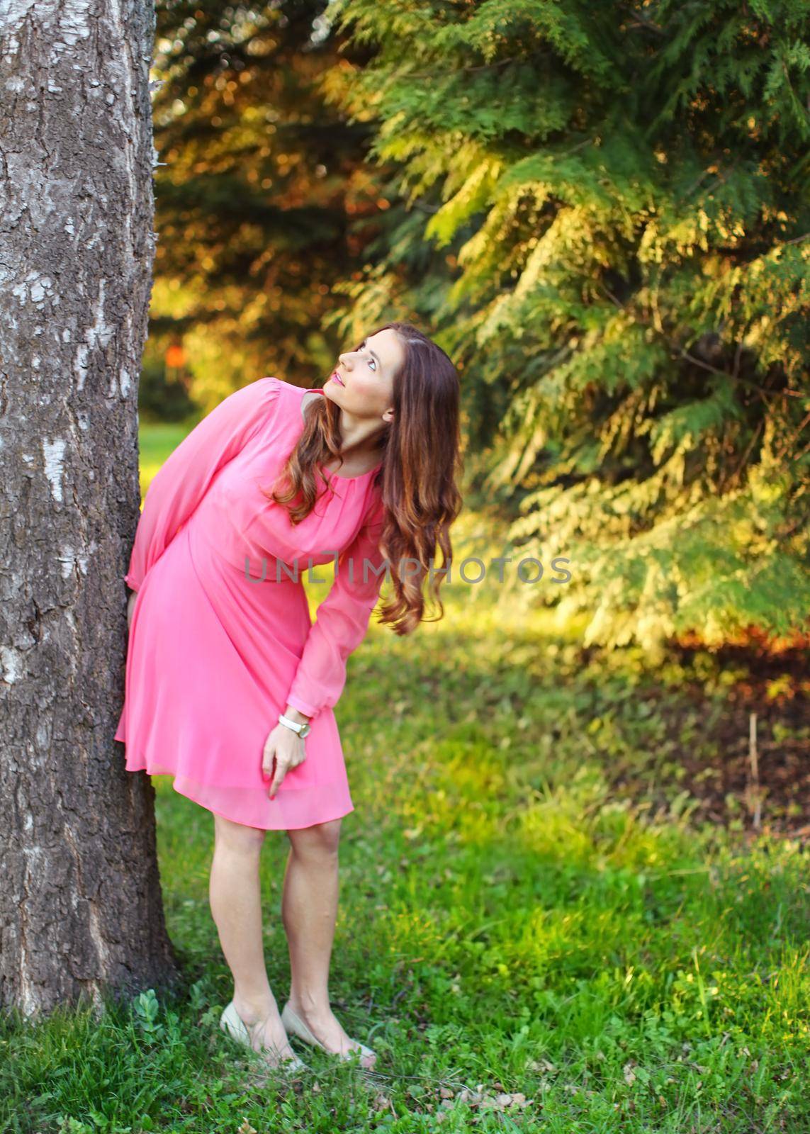 Young woman in pink dress, leaning to tree, looking up, with sunset light park in the background. by Ivanko