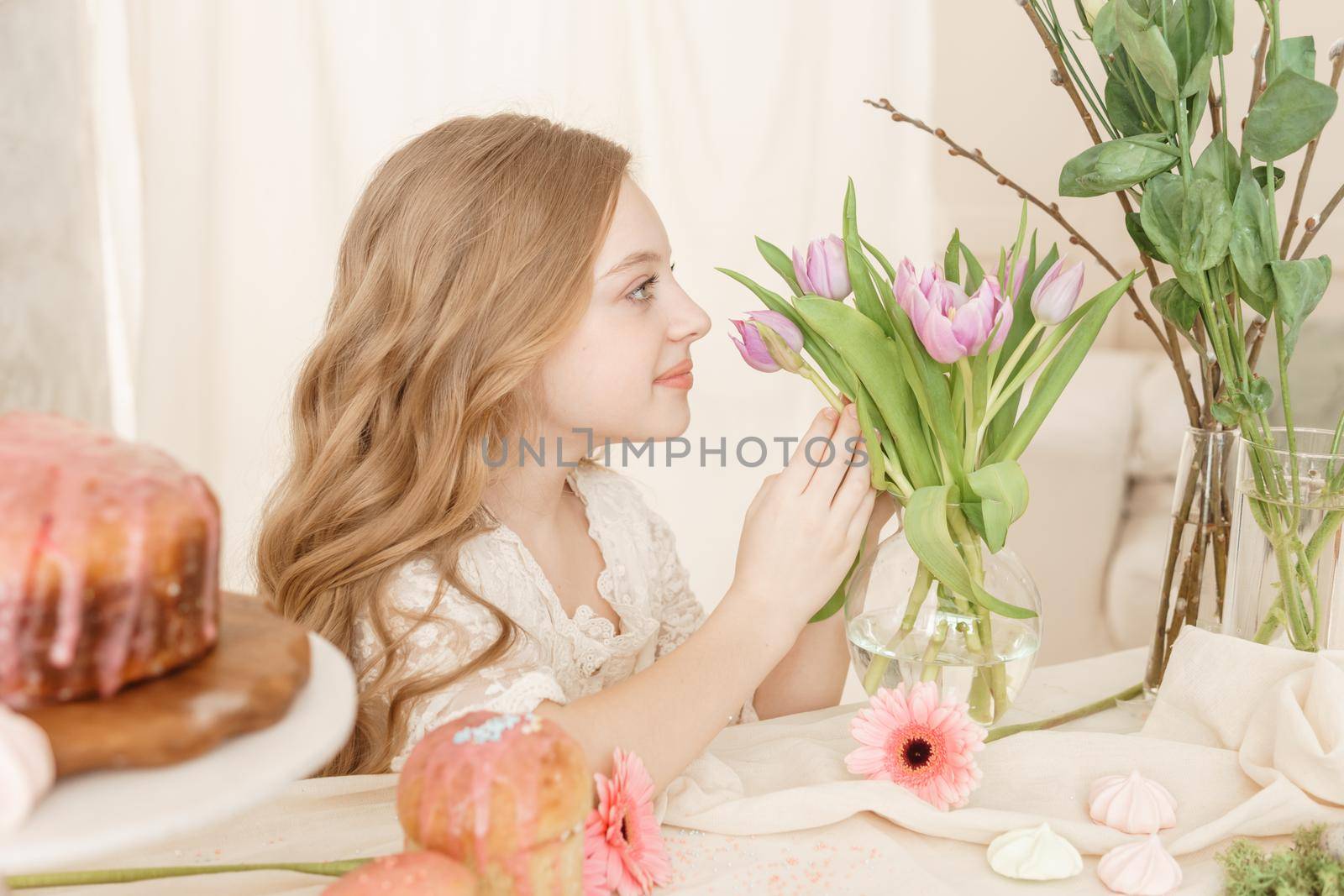 A girl with long hair in a light dress is sitting at the Easter table with cakes, spring flowers and quail eggs. Happy Easter celebration